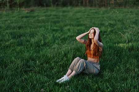 一位留着被风吹拂的长发的红发女性坐在户外公园的草地上微笑，落日的余晖照亮了她的脸庞。