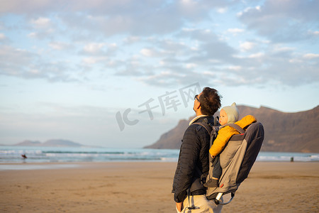 婴儿背包摄影照片_日落时，年轻的父亲在西班牙兰萨罗特岛 Famara 多风的沙滩上，一边享受纯净的大自然，一边背着他的婴儿男婴儿子背着背包，举起双手向天空。