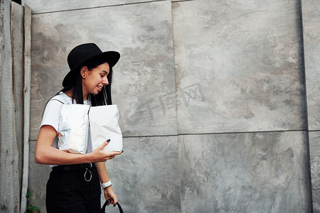 天气组件摄影照片_身穿黑帽和白衬衫的褐发女郎站在灰色墙壁附近，手里拿着带咖啡的包裹