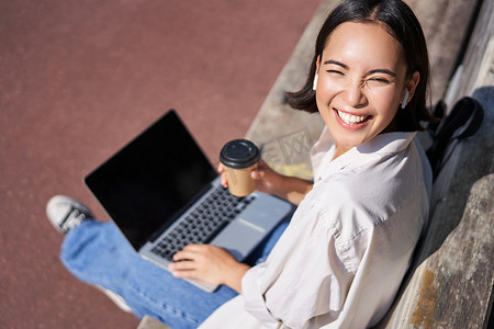 美丽的亚洲年轻女学生，带着笔记本电脑坐在公园的长椅上，喝外卖咖啡，听音乐，远程工作，在户外学习