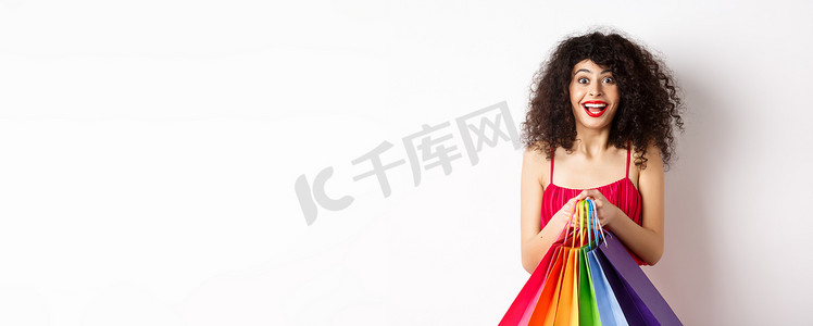 促销活动扁平风摄影照片_穿着时尚红裙、拿着购物袋微笑、在促销活动中购买、站在白色背景上的快乐年轻女性