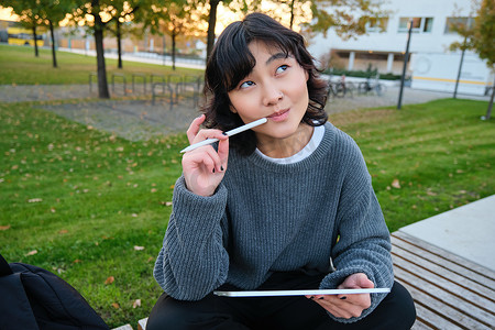 亚洲女性的肖像，公园里的学生，坐在长椅上，拿着数字平板电脑和一支笔，思考，一边思考，一边做笔记，在户外做作业