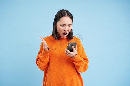 愤怒的韩国女孩对着手机骂骂咧咧，站在蓝色背景上，在智能手机上阅读新闻时看起来愤怒、愤怒