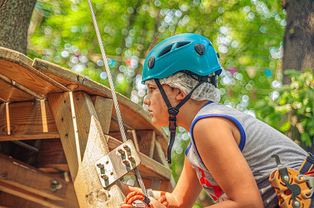 在森林绳索冒险公园的梯子上，一个戴着蓝色头盔和攀岩装备的十几岁男孩的侧面图