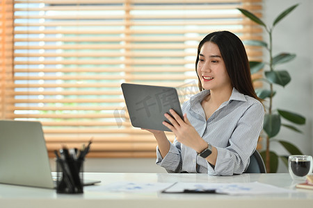 微笑的千禧一代女性坐在现代家庭办公室里阅读电子邮件或在数字平板电脑上检查日常工作