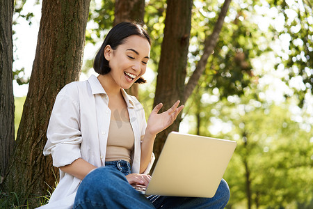 在阳光明媚的夏日，亚洲女性坐在树边的草地上，使用笔记本电脑，工作，远程做作业的肖像