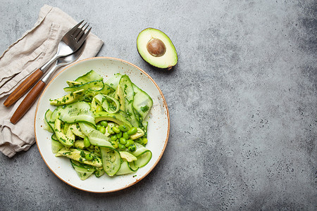 健康的纯素绿色鳄梨沙拉碗，配有切片黄瓜、毛豆、橄榄油和香草，陶瓷盘顶视图，灰色石头质朴的餐桌背景。