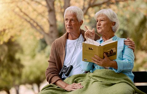 一对年长的夫妇在大自然中一起坐在公园的长椅上观鸟、读书和双筒望远镜。