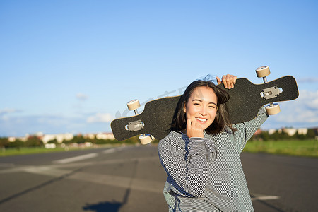 快乐而自由的亚洲女孩肩扛巡洋舰板，在空荡荡的路上走向镜头，在长板上滑冰，享受阳光明媚的天气