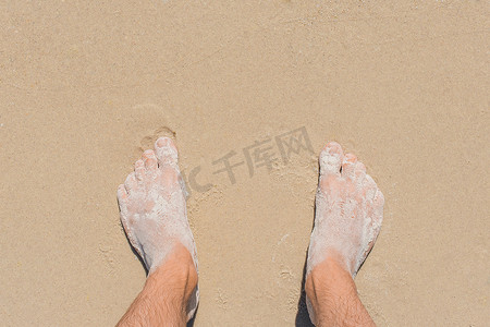 年轻人的腿站在沙滩上，被蓝色的海水冲刷，俯视