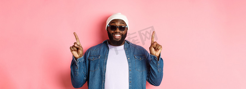 快乐的留着胡须的非洲裔美国人发布公告，指着手指微笑，站在粉红色背景中的帽子和太阳镜