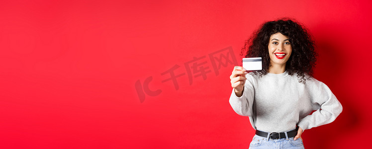 一头卷发、伸出手并展示塑料信用卡、推荐银行或购物优惠、红色背景的现代女性形象