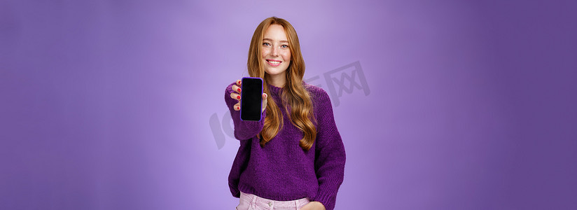 女孩在相机前展示智能手机屏幕，询问朋友的意见，面带乐观和快乐的表情，手牵着口袋，在紫色背景下推销手机或应用程序