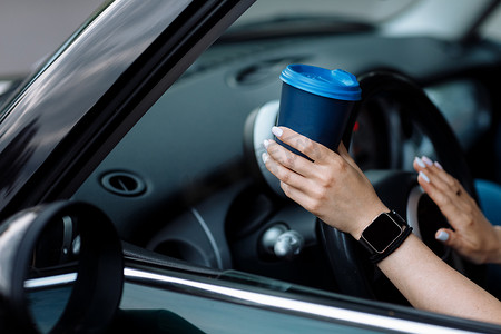 黑色车摄影照片_女人的手在车里拿着蓝纸杯咖啡的裁剪照片。