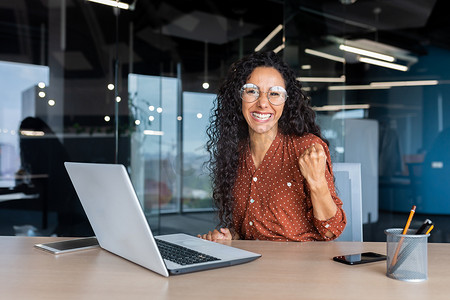 肖像成功的西班牙裔女商人办公室工作人员卷曲的头发微笑着看着相机在桌旁工作，笔记本电脑举起手势成功和胜利庆祝胜利。