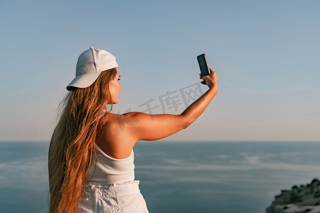 戴着帽子和背心的自拍女人制作自拍手机张贴照片社交网络户外海背景海滩人度假生活方式旅游概念。