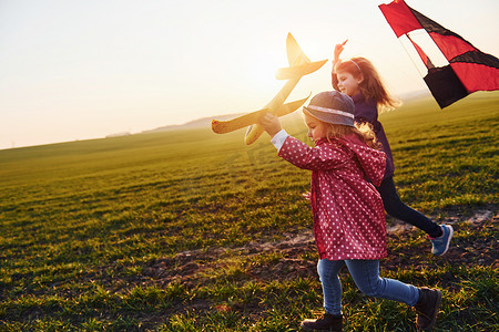 阳光明媚的白天，两个小女孩朋友在田野上玩风筝和玩具飞机