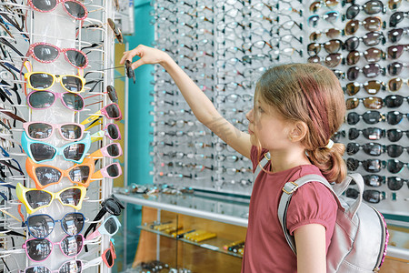 橱窗夏天摄影照片_女孩看和选择太阳镜，孩子靠近眼镜店的橱窗