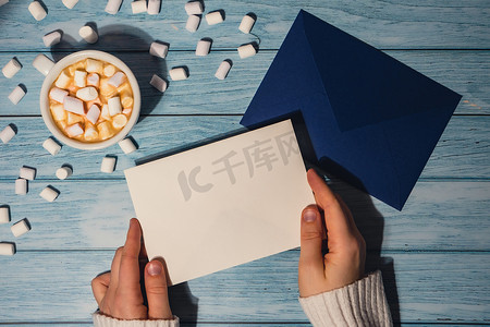 女手拿着空的问候语或邀请卡，用木制蓝色背景上的白色咖啡和棉花糖模拟蓝色信封。