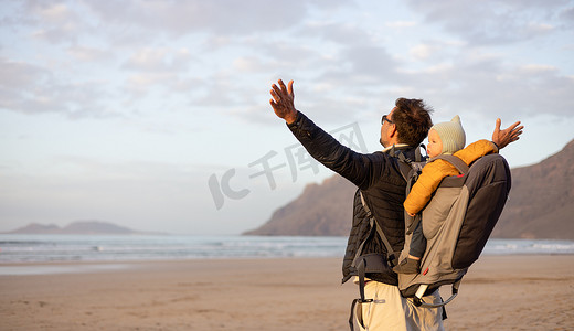 日落时，年轻的父亲在西班牙兰萨罗特岛 Famara 多风的沙滩上，一边享受纯净的大自然，一边背着他的婴儿男婴儿子背着背包，举起双手向天空。