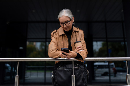 高龄女性摄影照片_自信的成熟女性自由职业者，戴着眼镜，头发灰白，在商务中心的背景下，手里拿着咖啡站着