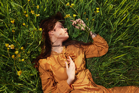 一个长着红色长发的平静女人躺在黄色花朵的绿色田野里，穿着橙色的连衣裙，闭着眼睛，把手放在脸旁，享受着平静和休养