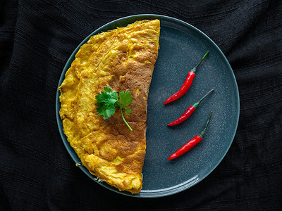 泰式煎蛋卷颜色为金黄色和红辣椒，放在绿色盘子上，鸡蛋顶部用香菜装饰，黑色背景