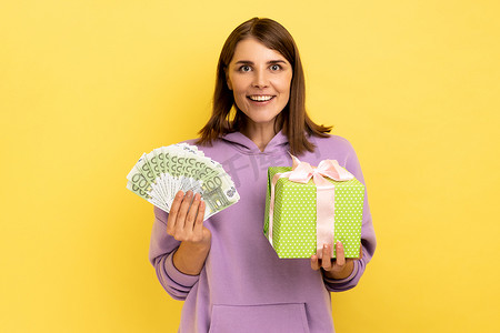 女人拿着包裹好的礼物盒和欧元纸币，用积极的表情看着相机。