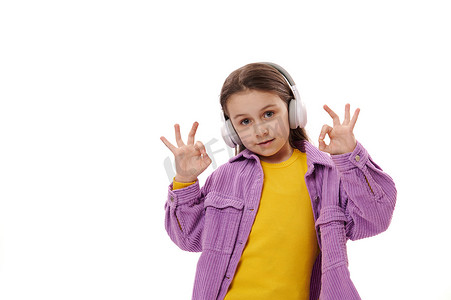 女生标志摄影照片_可爱的小女生喜欢用无线耳机听音乐，展示 OK 手势，可爱地微笑，看着镜头
