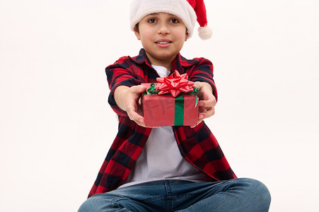 细节：一个戴着圣诞帽的男孩手里拿着红色蝴蝶结的圣诞礼物，在白色背景复制空间中被隔离。