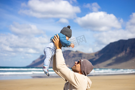 在西班牙兰萨罗特岛法马拉多风的沙滩上，父亲享受着纯净的大自然，抱着他的婴儿男婴儿子玩耍。