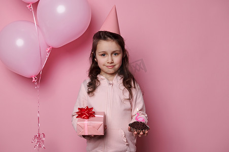 带蜡烛蛋糕摄影照片_穿着粉色衣服和派对帽的快乐小女孩，拿着可爱的生日礼物和带蜡烛的纸杯蛋糕