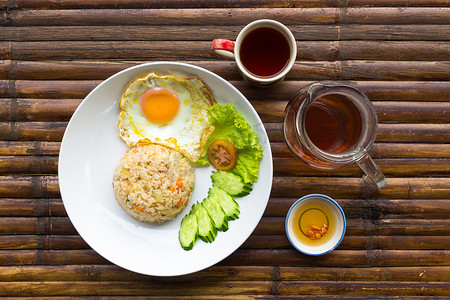 炒盘摄影照片_炒鸡蛋、米饭、白盘上的新鲜蔬菜、酱汁、水壶和茶杯，棕色竹桌上放着茶。