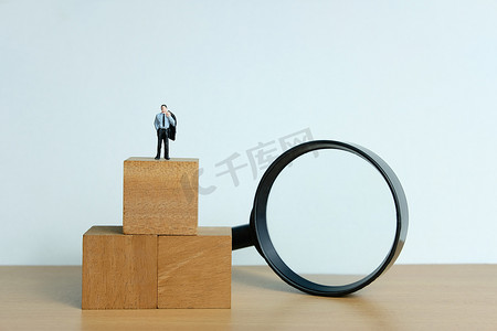 思考人物摄影照片_微型人物概念 — 一个绝望的商人站在木块上，用放大镜思考解决方案