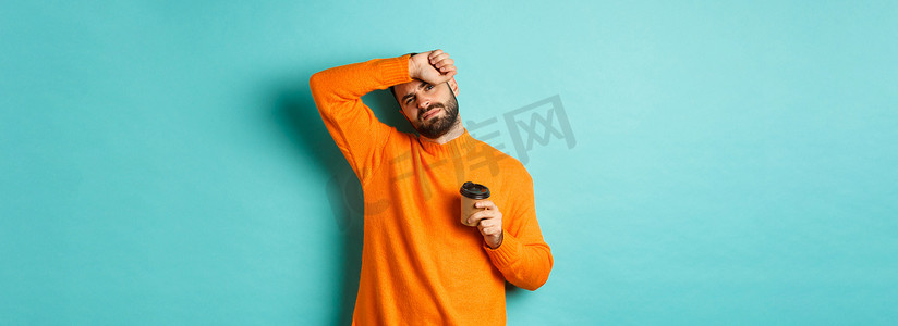 疲惫的成年男子下班休息喝咖啡，擦去额头上的汗水，看起来筋疲力尽，站在绿松石色背景上的橙色毛衣