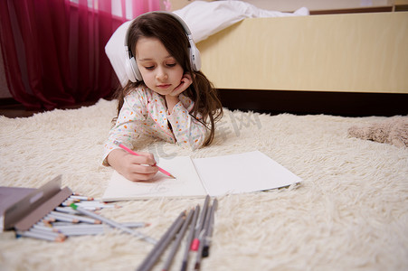 有才华的小女孩戴着耳机听音乐，用铅笔画画，躺在卧室的地毯上