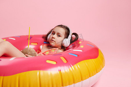 带无线耳机和粉红色充气游泳管上可可水的放松小女孩，与彩色背景隔离