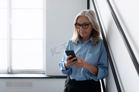 时尚的白人女商人在办公室靠墙看手机，白人中年老年女商人在工作概念