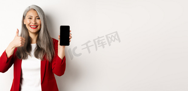 满意的亚洲老年女商人展示空白的智能手机屏幕和竖起大拇指，称赞在线促销或公司应用程序，站在白色背景上