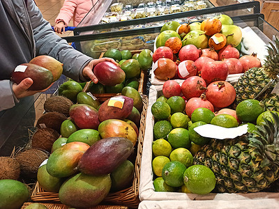 超市水果柜台摄影照片_特写顾客拿着成熟的有机芒果，站在杂货店柳条篮里放着新鲜水果的柜台旁