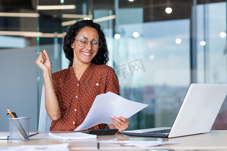 女商人庆祝胜利和成功成就，西班牙裔女性在文书工作背后阅读带有积极结果报告的文件，女性在办公室坐在桌旁使用笔记本电脑工作