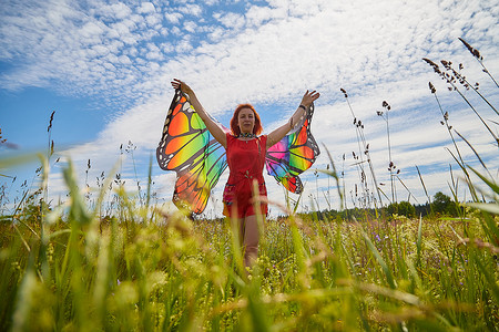 夏日玩乐摄影照片_有红头发和蝴蝶翅膀的成年女孩在草地或草地上玩乐，在夏日阳光明媚的日子里花