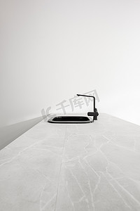 水槽广告摄影照片_带时尚水龙头的黑色金属厨房水槽位于浅色背景的大型白色石头厨房岛上。