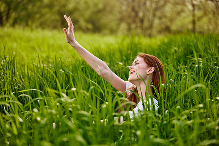 快乐、心满意足的女人坐在高高的绿草上，高高举起双臂享受愉快的一天