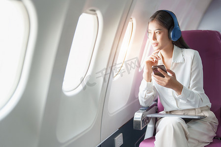 坐飞机摄影照片_年轻的亚洲美女乘客坐在商务舱豪华飞机上，同时戴着无线耳机，在飞行期间使用智能手机放松