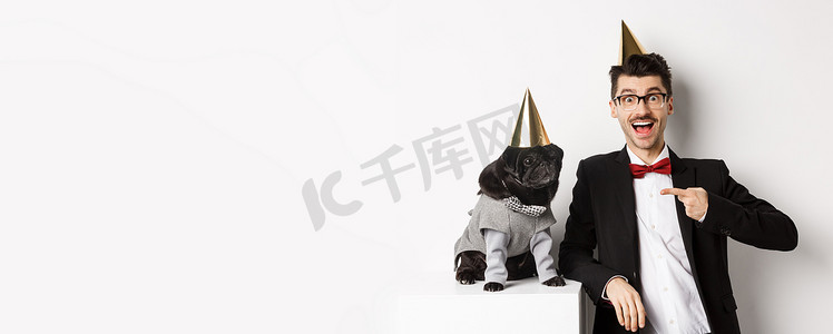 快乐的男人和可爱的黑哈巴狗穿着派对锥和西装，狗主人庆祝宠物生日，站在白色背景上