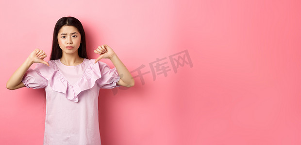 持怀疑态度和失望的亚洲女性不情愿地笑着竖起大拇指，不喜欢产品，表现出负面情绪，站在粉红色背景中