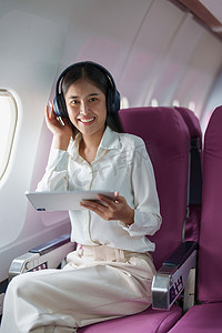 孤寂摄影照片_年轻的亚洲美女乘客坐在商务舱豪华飞机上，同时戴着无线耳机在飞行期间放松和使用平板电脑
