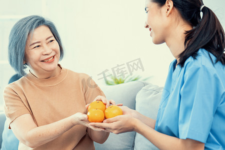 心满意足摄影照片_一位年轻的护理人员在家中将橙子递给心满意足的老年患者。