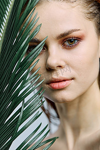 一个美丽的女人站着，手里拿着一片热带棕榈叶，把它带到她的脸上，看着镜头。